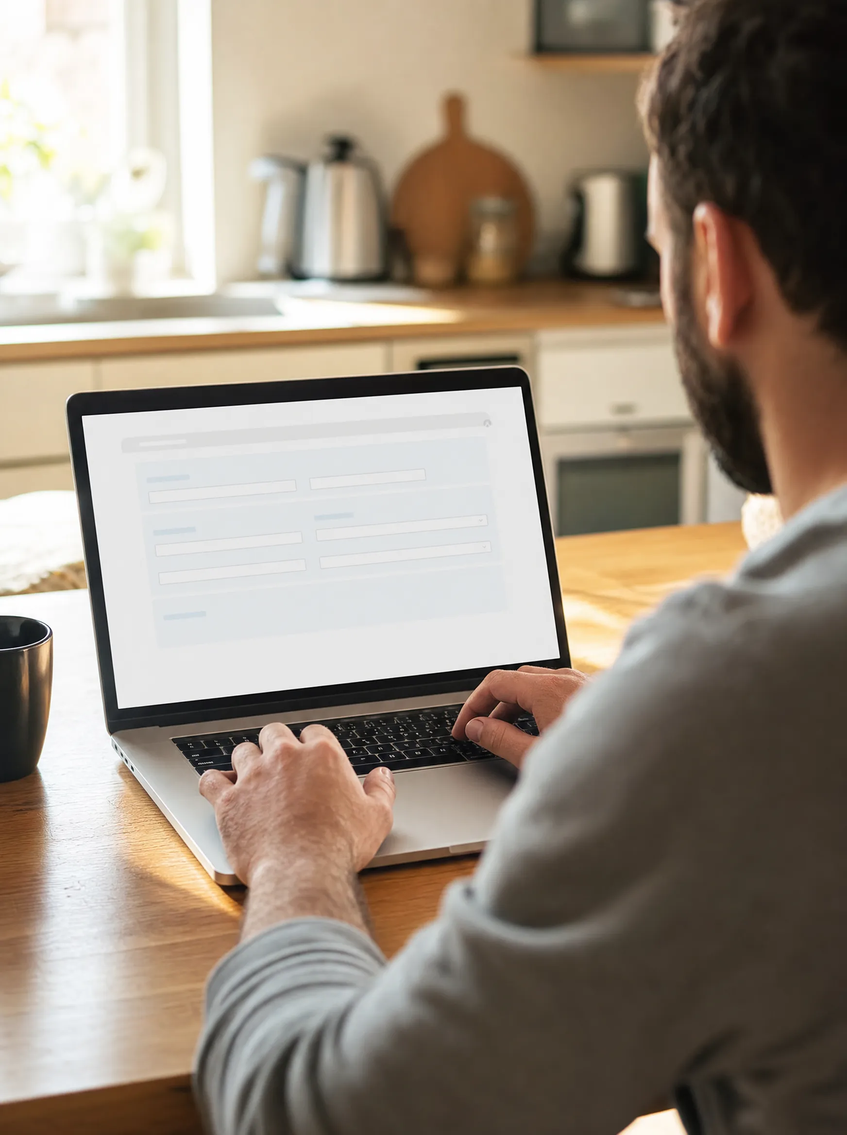 Over-the-shoulder view of a person filling out a web form on their laptop at a sunlit kitchen table, the first step of the application process.