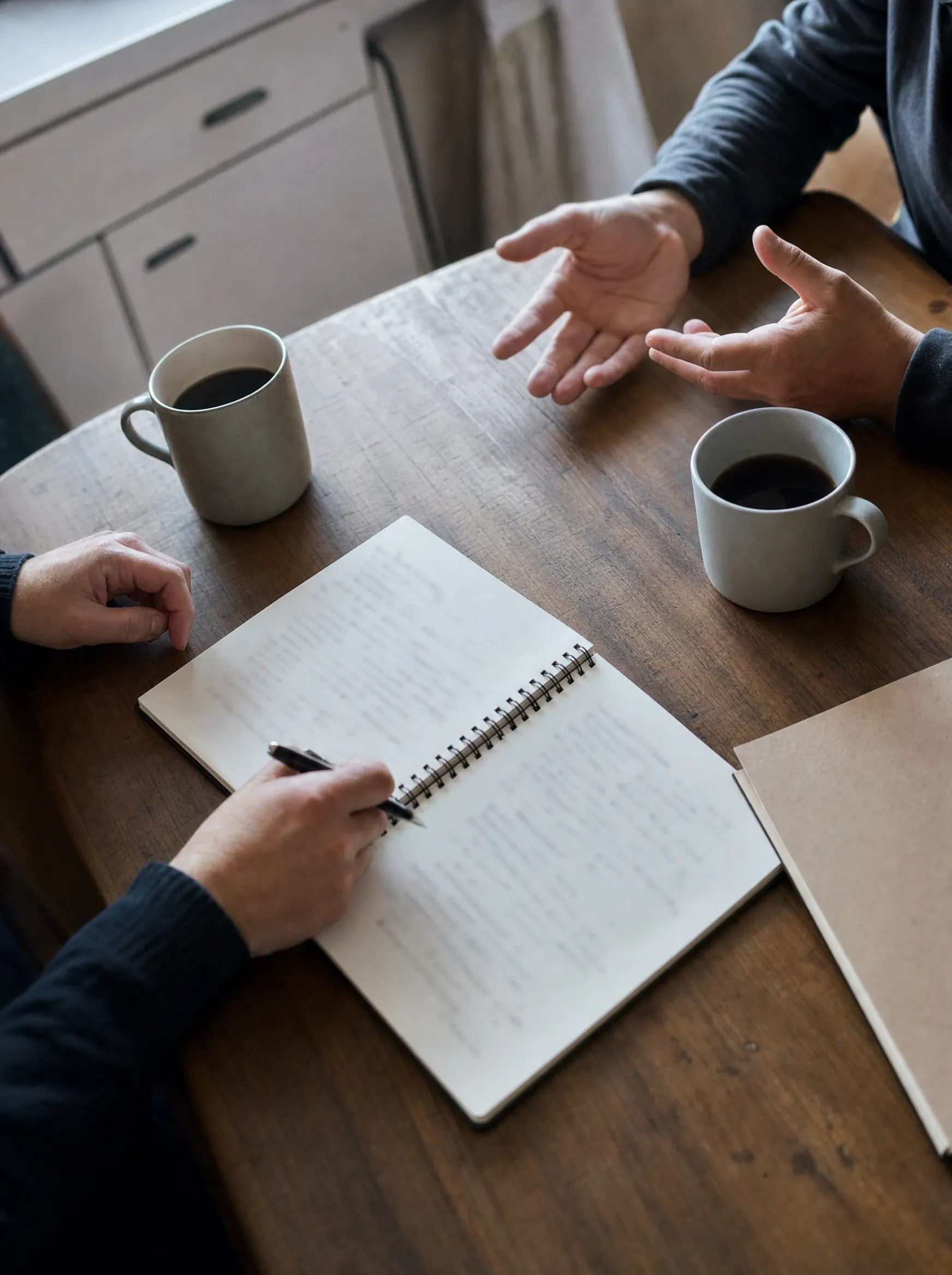 Overhead view of two people working through a notebook and folder on a kitchen table with coffee mugs.